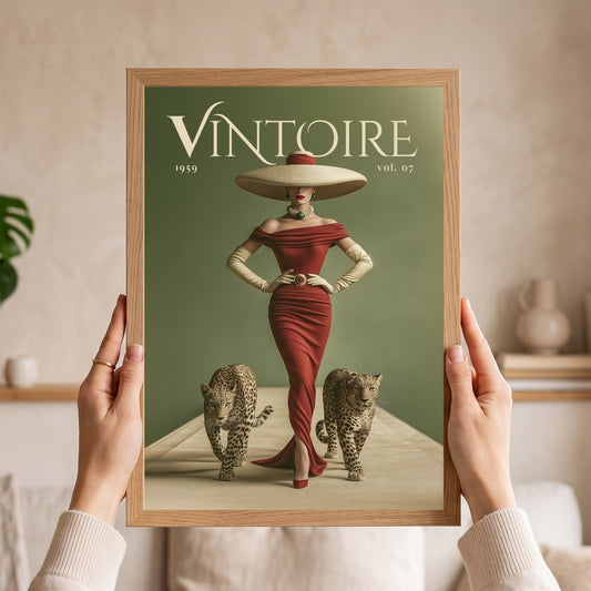 A person holds a framed vintage-style art print featuring the word 'VINTOIRE' at the top, a woman in a red dress and large hat, and two cats on a muted green background. The scene is indoors with a softly blurred neutral home setting behind the hands and frame.