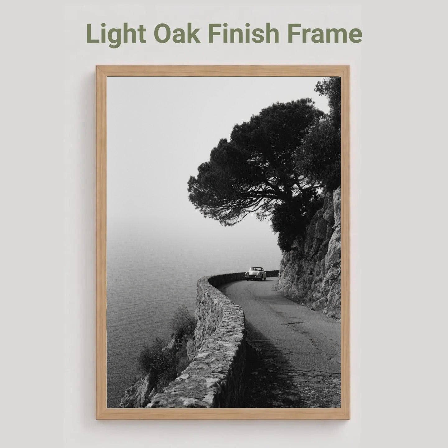 A framed black and white coastal road print with a tree and cliff, shown against a light gray background with the text 'Light Oak Finish Frame' above it.