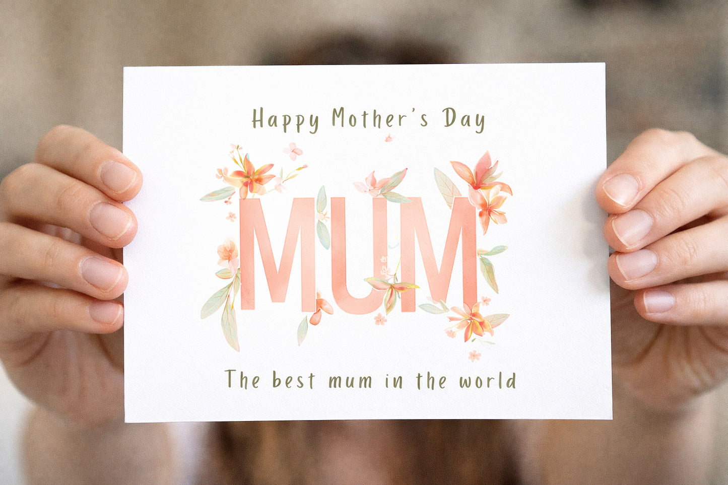 Hand holding a Mother's Day card with 'Mum' and floral decorations. Mother’s Day greeting card with pastel floral watercolor design, large “MUM” text, and wording “Happy Mother’s Day” and “The best mum in the world”.