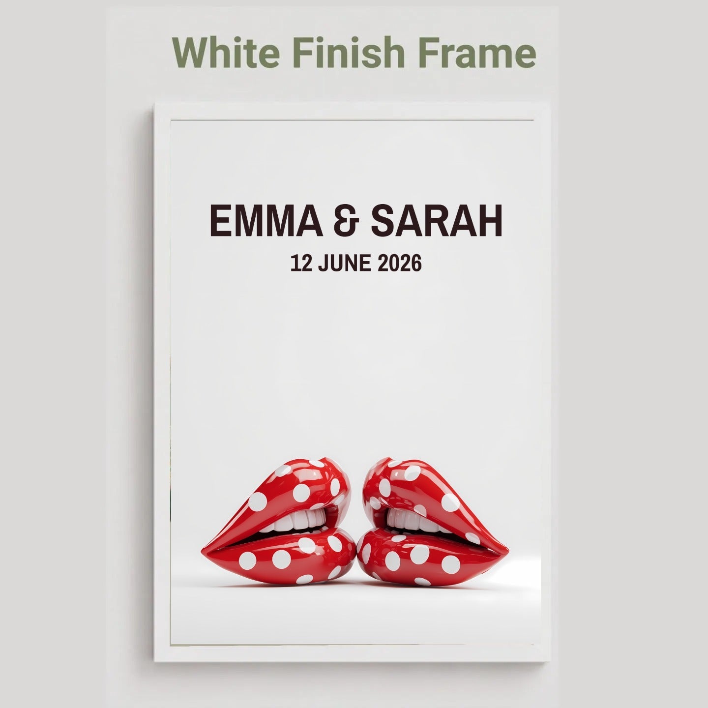 A framed custom wedding print with a white finish frame, featuring the text 'EMMA & SARAH' and '12 JUNE 2026' above a graphic of two red lips with white polka dots, on a light gray background.