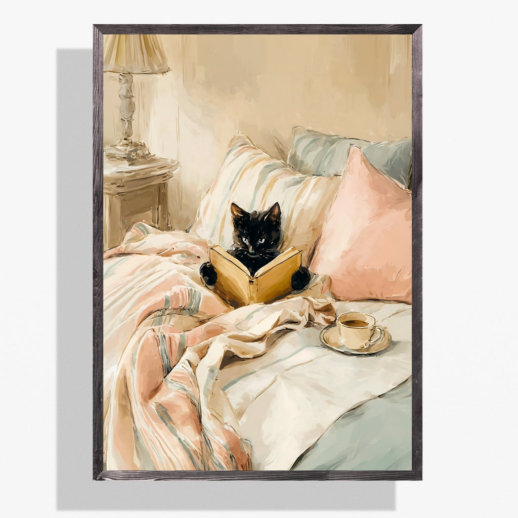 Black-framed poster of a black cat reading a book in bed among blush pink and cream pillows, with a cup of coffee or tea on a saucer.