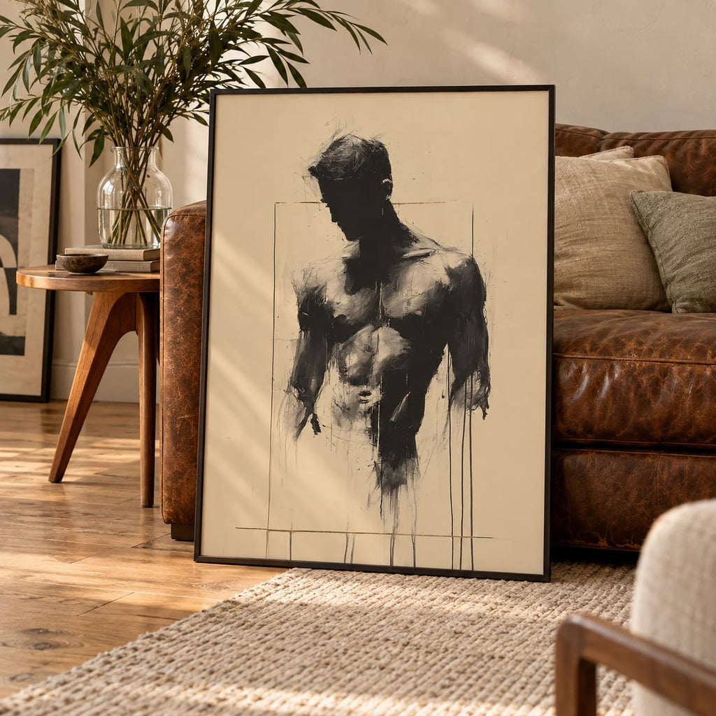 Bare Masculinity male figure print in a black frame leaning against a vintage brown leather sofa beside a wooden side table and a plant.