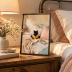 Framed black cat reading print on a wooden bedside table beside a glowing lamp and flowers, in blush pink and cream tones.