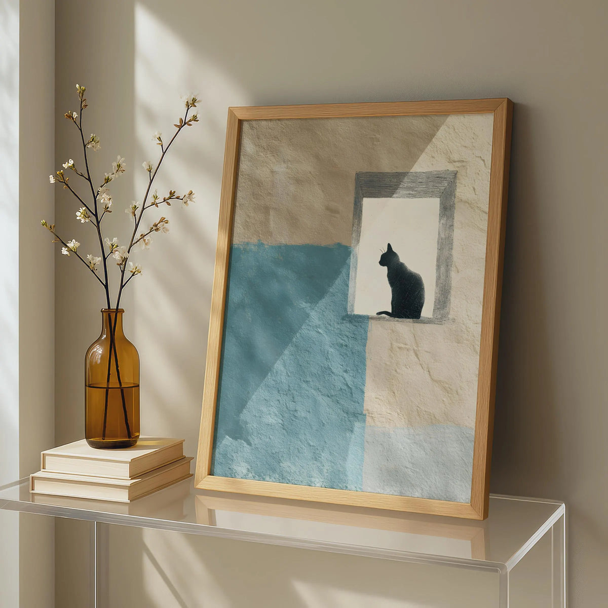 A framed art print leaning on a glass table against a beige wall, featuring a black cat silhouette in a window over a blue and cream textured background. A brown glass vase with white blossoms and stacked books sit beside it.