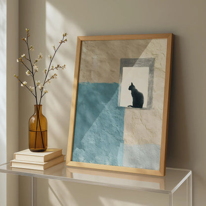 A framed art print leaning on a glass table against a beige wall, featuring a black cat silhouette in a window over a blue and cream textured background. A brown glass vase with white blossoms and stacked books sit beside it.