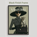 A framed art print featuring a vintage-style fashion illustration with the text 'Black Finish Frame' above it. The artwork shows a woman in a black wide-brim hat and black-and-white polka dot dress holding a small spotted kitten over a black handbag, with the magazine-style title 'VINTOIRE' across the top.