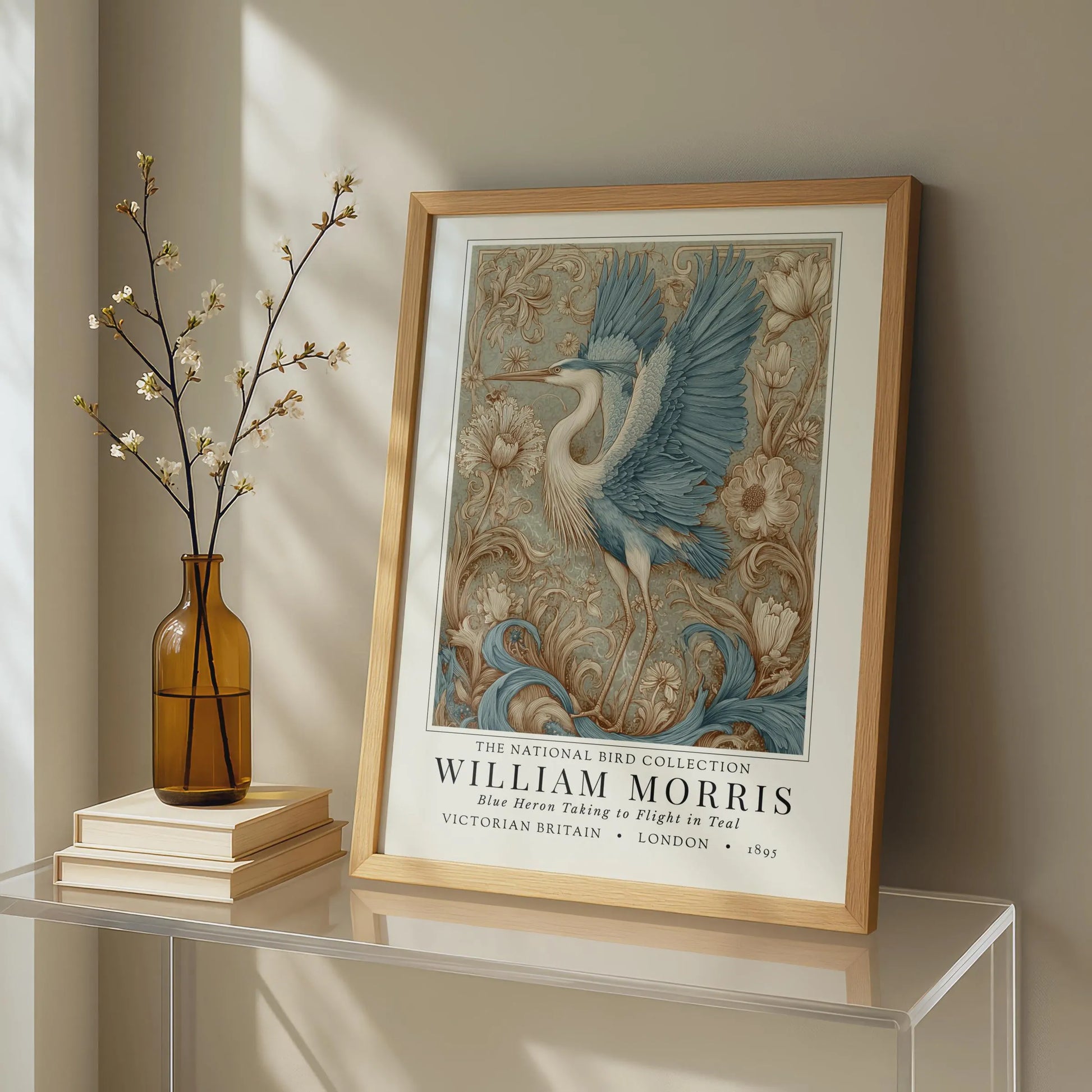Framed artwork of a bird with decorative elements on a shelf next to a vase and books. William Morris style exhibition poster featuring a blue heron taking to flight, surrounded by ornate acanthus scrolls, dahlias and botanical foliage in teal, gold and antique cream — Arts & Crafts wall art print