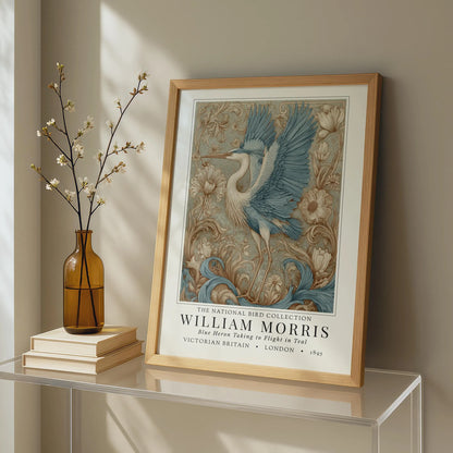 Framed artwork of a bird with decorative elements on a shelf next to a vase and books. William Morris style exhibition poster featuring a blue heron taking to flight, surrounded by ornate acanthus scrolls, dahlias and botanical foliage in teal, gold and antique cream — Arts & Crafts wall art print