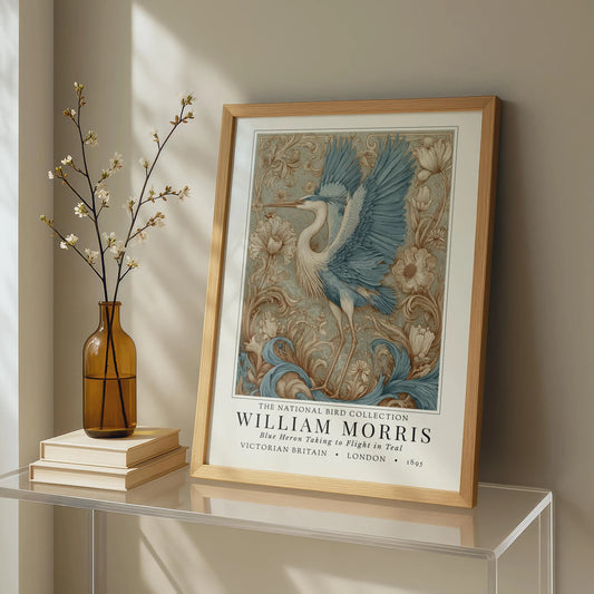 Framed artwork of a bird with decorative elements on a shelf next to a vase and books. William Morris style exhibition poster featuring a blue heron taking to flight, surrounded by ornate acanthus scrolls, dahlias and botanical foliage in teal, gold and antique cream — Arts & Crafts wall art print