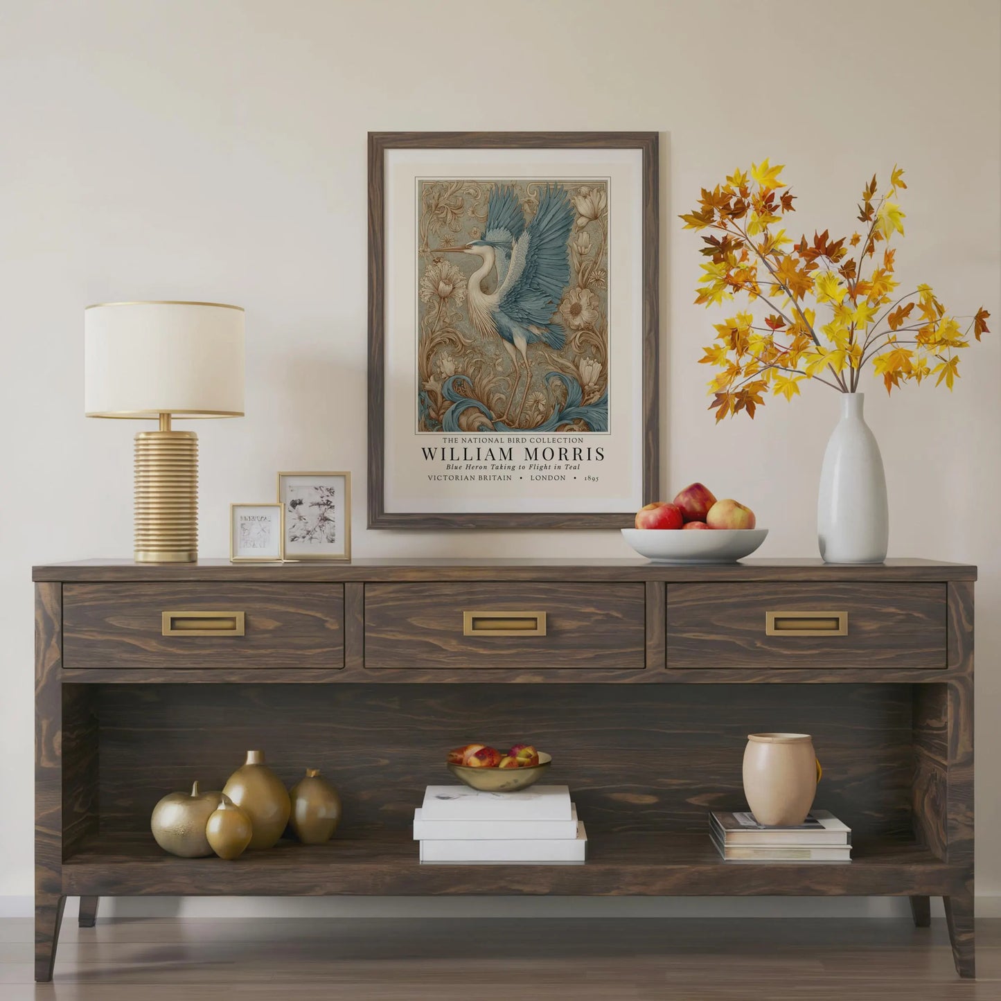 Wooden console table with decorative items including a lamp, framed artwork, and vases. William Morris style exhibition poster featuring a blue heron taking to flight, surrounded by ornate acanthus scrolls, dahlias and botanical foliage in teal, gold and antique cream — Arts & Crafts wall art print