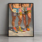 A framed art print leaning against a gray wall on a light wood floor. The print shows two cropped male figures from the waist down wearing colorful patchwork shorts and brightly colored athletic sneakers.