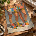 Unframed Cool Boys poster lying on a sunlit wooden desk with a plant, books and a cup of coffee, showing two men in pastel patchwork shorts and colourful retro trainers.