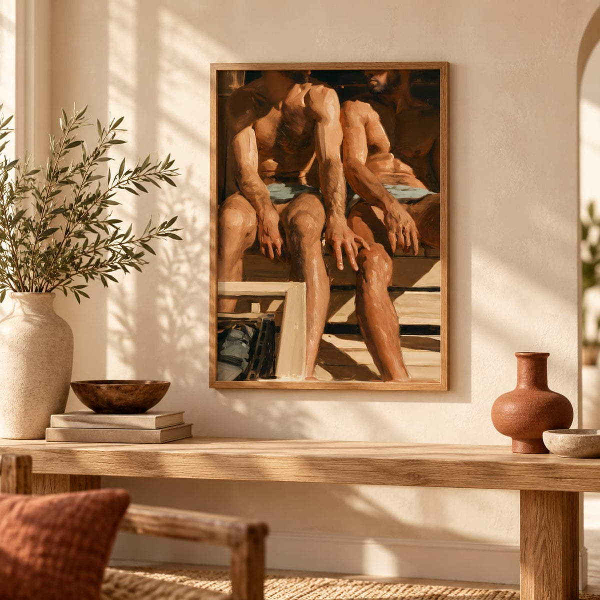 A framed art print depicting two muscular men sitting on a bench, styled on a beige wall above a wooden console table in a warm, sunlit room.