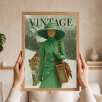 A person holds a framed vintage-style art print featuring a woman in a green outfit and hat walking with a leopard in the snow. The print has the word "VINTAGE" at the top and is shown against a softly lit indoor background.