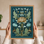 A framed botanical art print with a floral design in teal, blue, cream, and gold tones, held by two hands in front of a softly blurred indoor background.