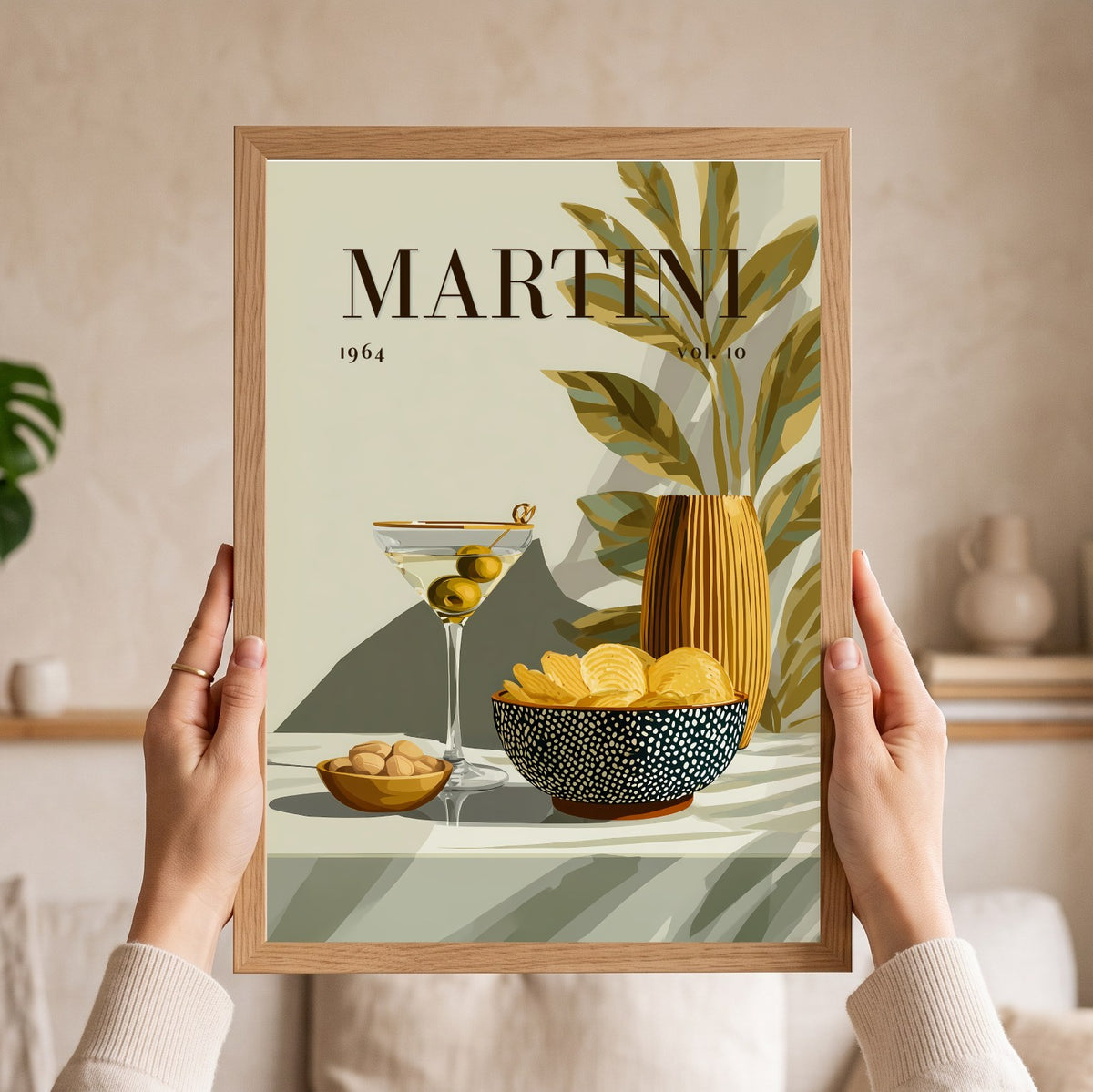 A person holds a framed art print featuring a retro-style Martini poster with a martini glass, olives, chips, and a vase. The print has a light wood frame and is held by two hands against a softly blurred beige living room background.
