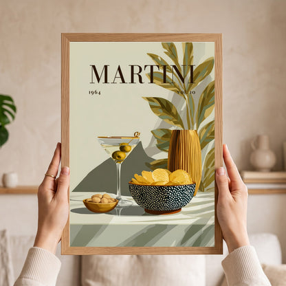 A person holds a framed art print featuring a retro-style Martini poster with a martini glass, olives, chips, and a vase. The print has a light wood frame and is held by two hands against a softly blurred beige living room background.