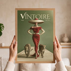 A person holds a framed vintage-style art print featuring the word 'VINTOIRE' at the top, a woman in a red dress and large hat, and two cats on a muted green background. The scene is indoors with a softly blurred neutral home setting behind the hands and frame.