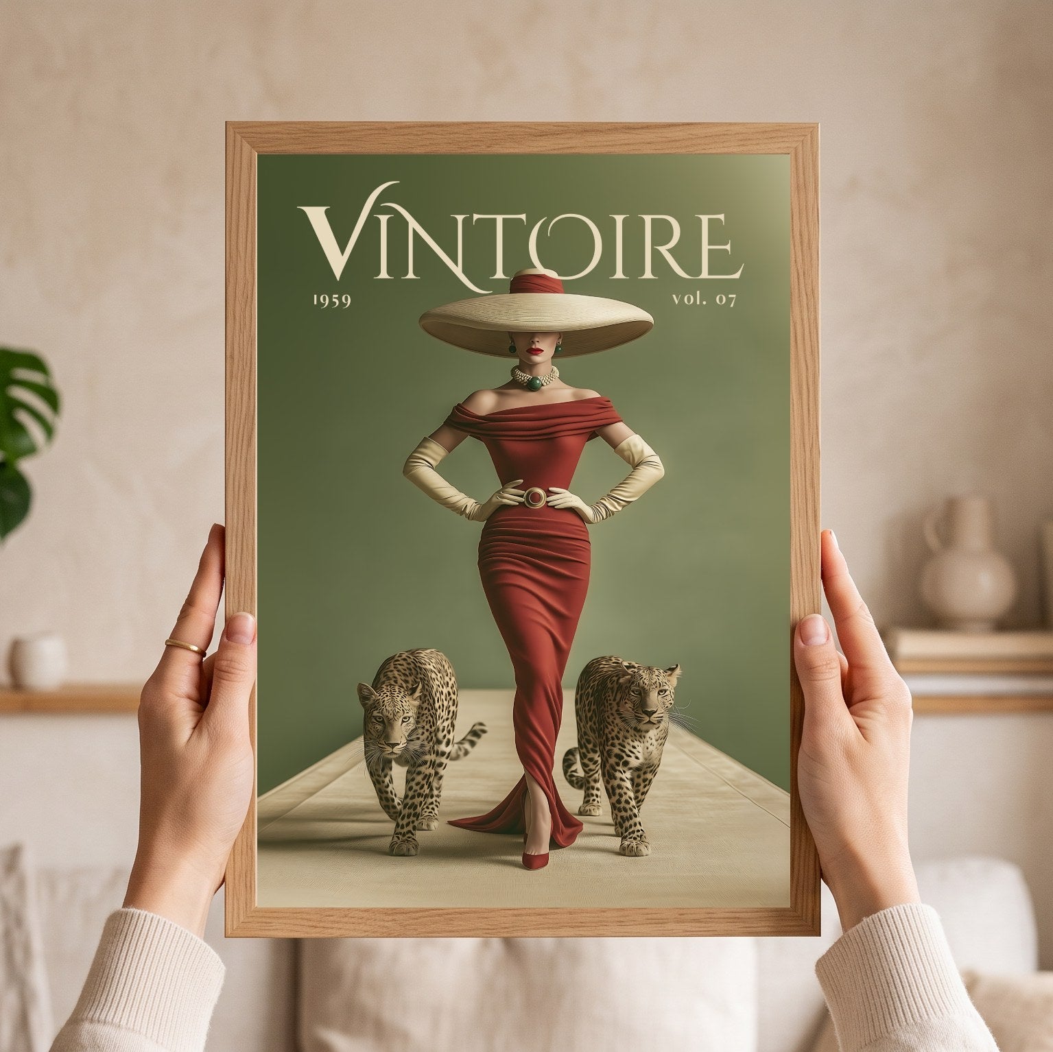 A person holds a framed vintage-style art print featuring the word 'VINTOIRE' at the top, a woman in a red dress and large hat, and two cats on a muted green background. The scene is indoors with a softly blurred neutral home setting behind the hands and frame.
