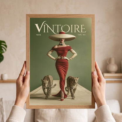 A person holds a framed vintage-style art print featuring the word 'VINTOIRE' at the top, a woman in a red dress and large hat, and two cats on a muted green background. The scene is indoors with a softly blurred neutral home setting behind the hands and frame.