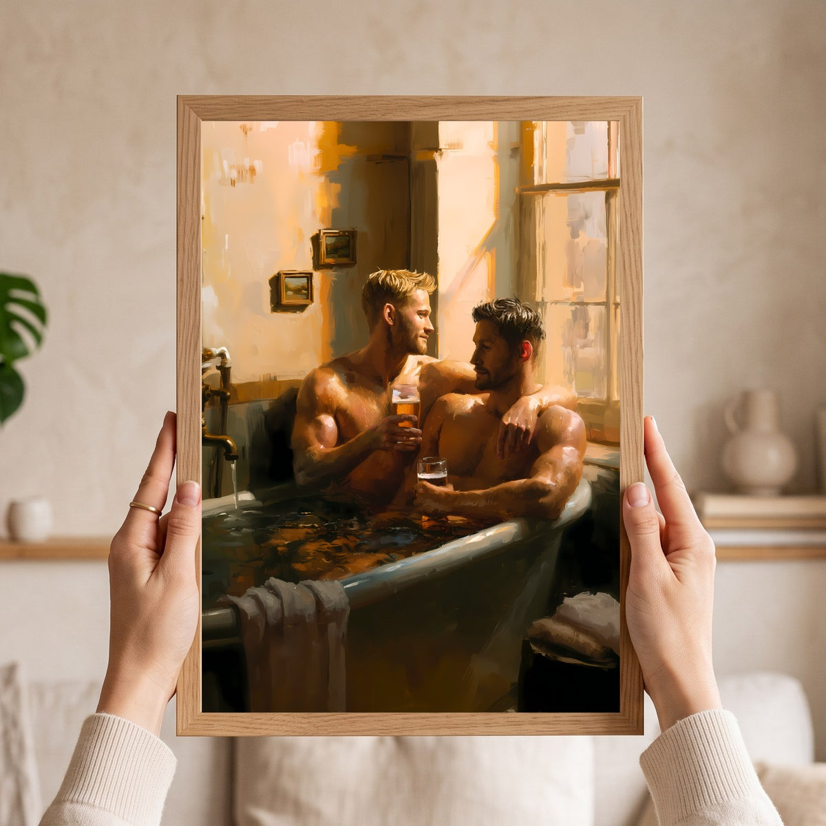 A person holds a framed art print featuring a warm-toned painting of two men sitting in a bathtub. The print has a light natural wood frame, and the background shows a softly blurred beige interior with a plant and shelf.