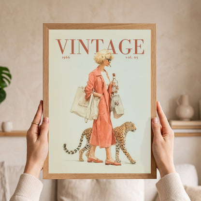 A person holds a framed vintage-style art print featuring a woman in a peach dress walking beside a cheetah, with the word “VINTAGE” at the top. The print is shown in a light wood frame against a softly lit indoor background.