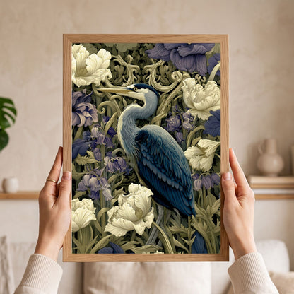 A person holds a framed art print featuring a detailed illustration of a blue heron surrounded by white flowers and green foliage, set against a softly blurred beige indoor background.