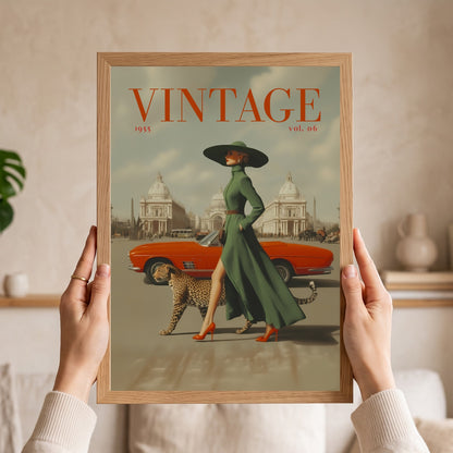A person holds a framed vintage-style art print featuring a woman in a long green dress and black hat walking beside an orange convertible car and a leopard, with the word "VINTAGE" at the top. The print is shown against a softly lit indoor background with a plant and shelf visible.