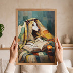 A person holds a framed art print featuring a stylized painting of a greyhound dog wrapped in a blanket and reading a book. The hands holding the frame are visible against a softly blurred indoor background with shelves and decor.