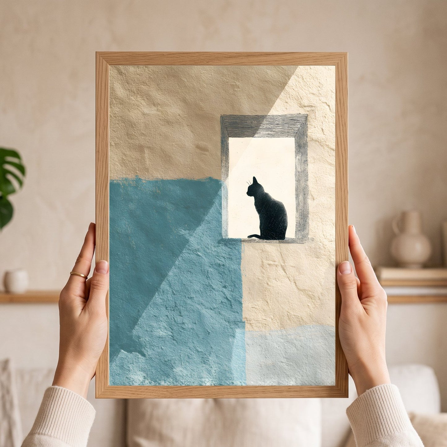 A person holds a framed art print featuring a black cat silhouette sitting in a window-like frame against a textured beige and light blue background. The scene is set in a softly lit, modern living space with neutral decor.