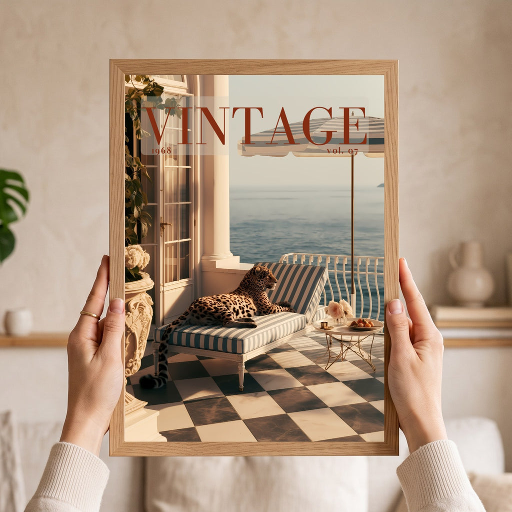 A person holds a framed art print featuring a vintage magazine cover design with a leopard lounging on a striped chaise lounge by the sea. The print is housed in a light natural wood frame, and the person's hands and arms are visible holding it against a softly blurred beige interior background.