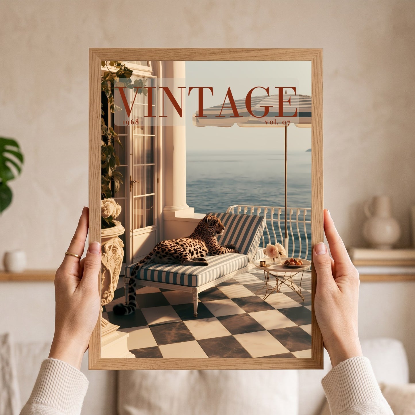 A person holds a framed art print featuring a vintage magazine cover design with a leopard lounging on a striped chaise lounge by the sea. The print is housed in a light natural wood frame, and the person's hands and arms are visible holding it against a softly blurred beige interior background.