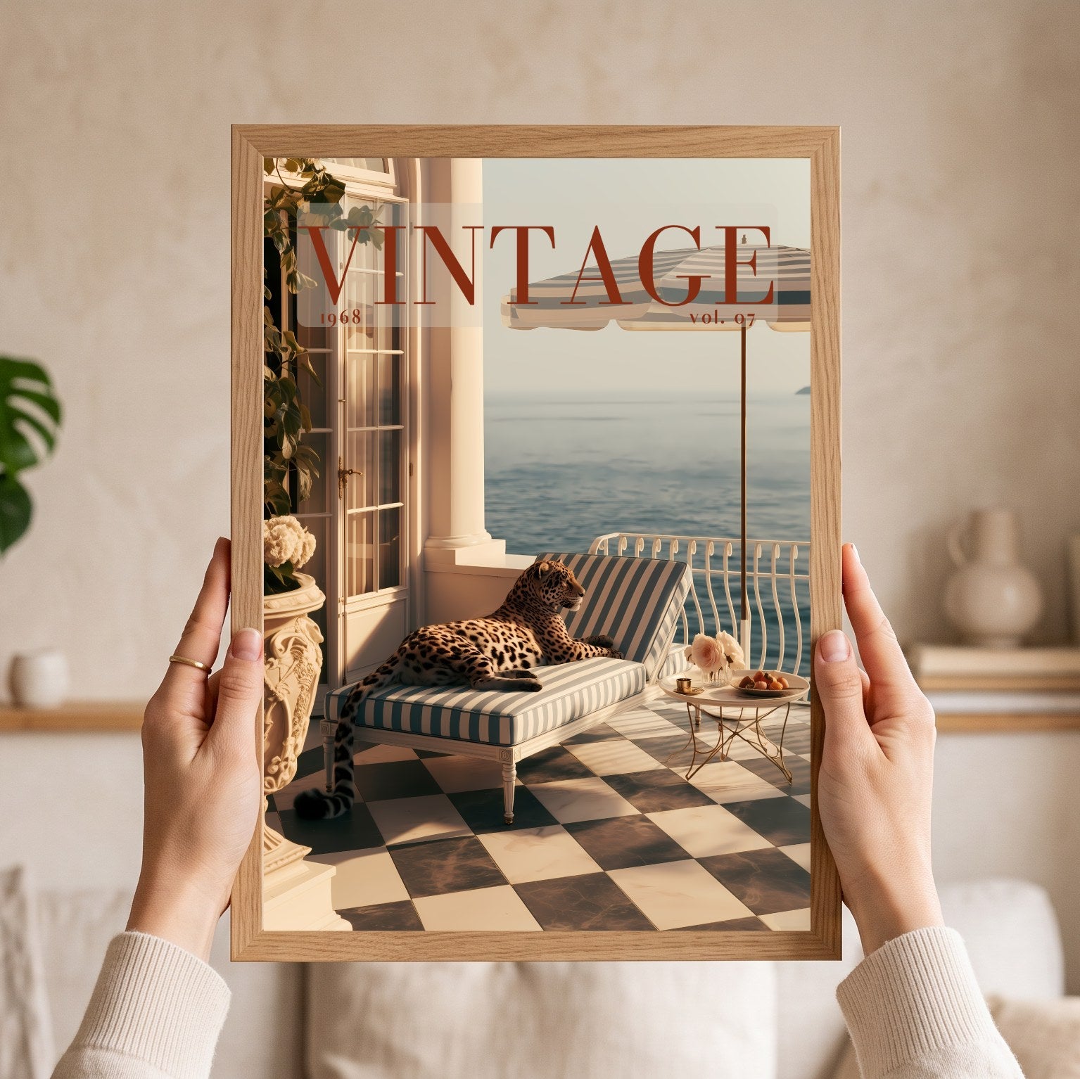 A person holds a framed art print featuring a vintage magazine cover design with a leopard lounging on a striped chaise lounge by the sea. The print is housed in a light natural wood frame, and the person's hands and arms are visible holding it against a softly blurred beige interior background.