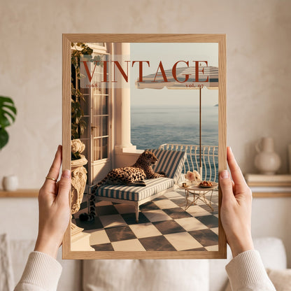 A person holds a framed art print featuring a vintage magazine cover design with a leopard lounging on a striped chaise lounge by the sea. The print is housed in a light natural wood frame, and the person's hands and arms are visible holding it against a softly blurred beige interior background.