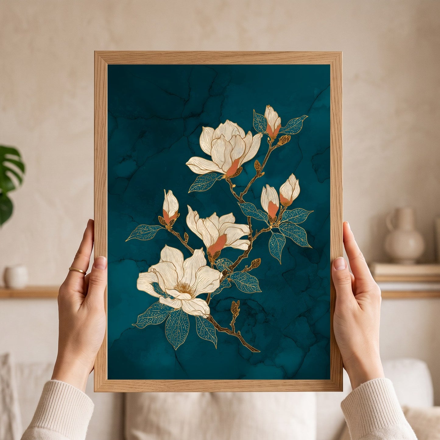 A person holds a framed art print featuring white magnolia flowers with gold accents on a dark teal marbled background. The print is housed in a light oak wood frame, and the background shows a softly blurred beige living room interior.