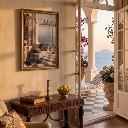 A framed vintage-style art print featuring a balcony scene with the word 'VINTAGE' hangs on a beige wall in a sunlit interior. Below the frame is a dark wood console table with books, a candlestick, and a bowl of lemons.