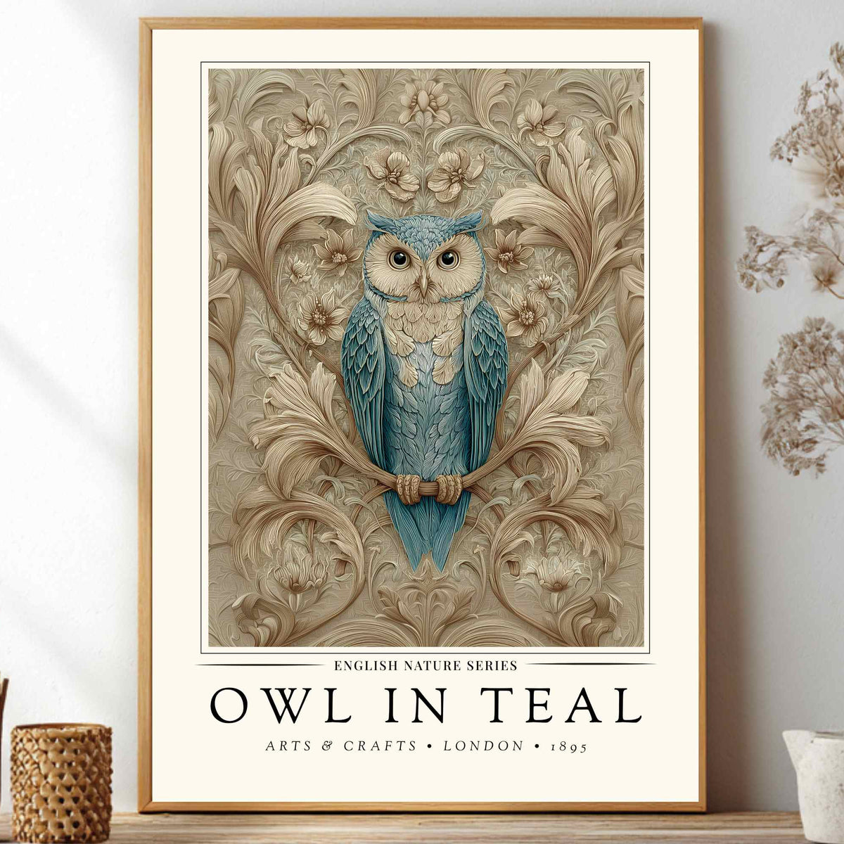 A framed art print featuring a blue owl perched on a branch against an ornate beige floral background, with the text 'OWL IN TEAL' and 'ARTS & CRAFTS • LONDON • 1893' below. The print is displayed leaning against a white wall on a shelf, surrounded by decorative dried flowers and small vessels.