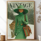 A framed vintage-style poster or magazine cover print leaning against a wall on a shelf, showing a woman in a green outfit and hat with a leopard in the snow. The print is surrounded by home decor props including dried stems in a glass vase, a small white pot, and woven containers.