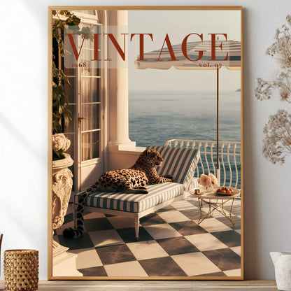 A framed vintage-style poster leaning against a white wall in a styled interior. The poster shows a balcony scene with a striped lounge chair, leopard-print pillow, ocean view, and large 'VINTAGE' text at the top.