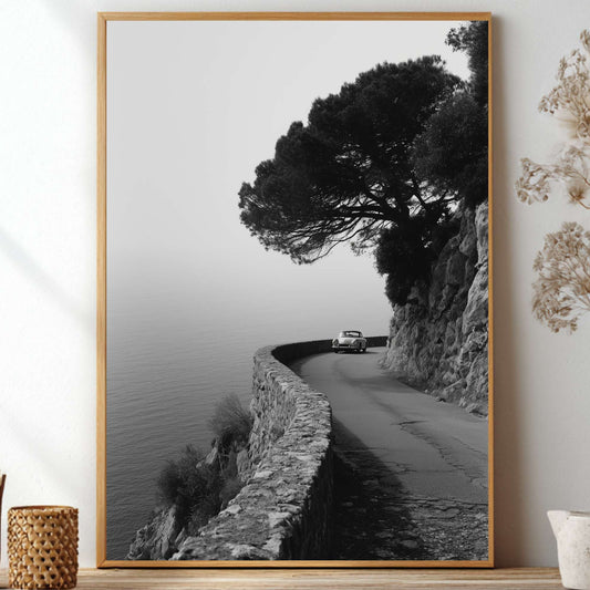 A vertical framed black and white landscape photograph of a winding coastal road with a lone tree, displayed on a shelf in a styled interior setting with dried flowers and decorative vases.