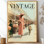 A framed vintage-style art print featuring a woman in a pink dress and hat walking a leopard on a city street, with large 'VINTAGE' text at the top. The framed print is displayed on a shelf against a light wall, surrounded by decorative dried flowers and small vases.