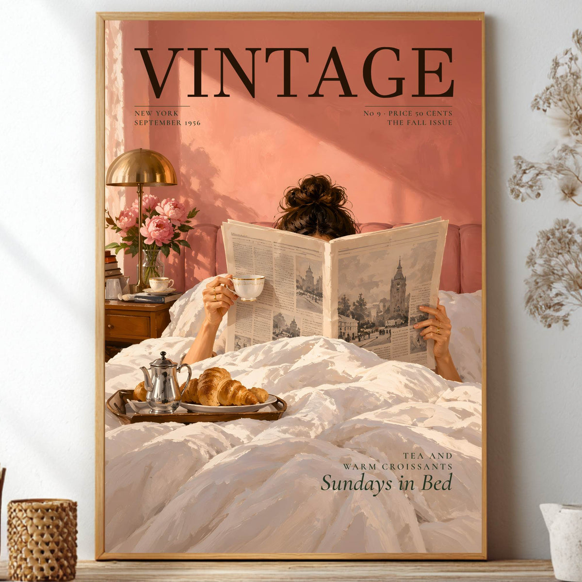 A framed vintage-style poster or print leaning on a shelf, showing a person reading a newspaper in bed with a cup and tea, against a pink background. The poster is surrounded by home decor props including dried flowers, a lamp, and small vases.