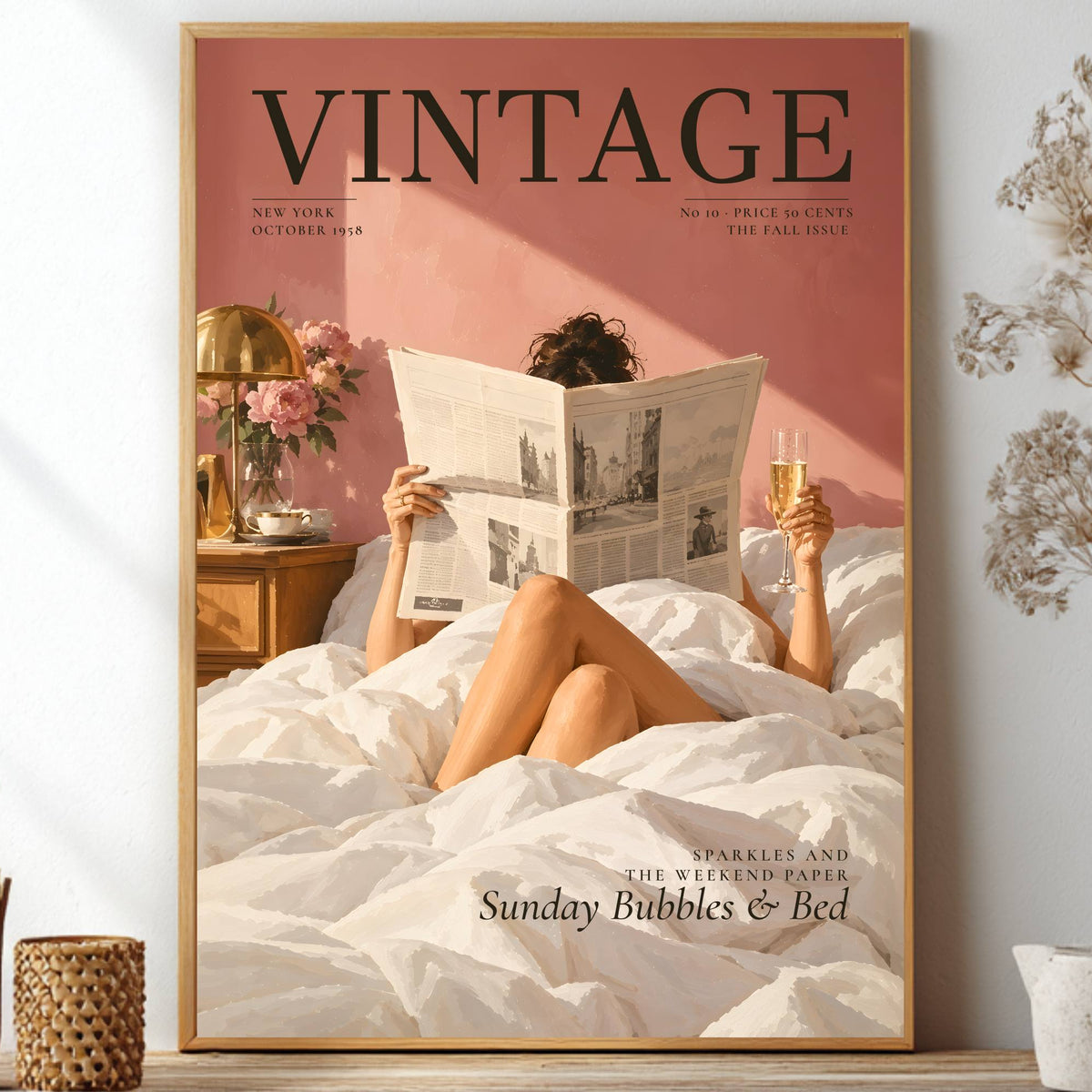 A framed vintage-style wall art print featuring a woman reading a newspaper in bed, styled on a shelf between decorative dried flowers and small vases. The poster has a dusty pink background, black serif text reading 'VINTAGE,' and white bedding.