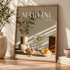 A framed art print leaning against a wall on a wooden floor, featuring a minimalist Martini glass, bowls of snacks, and the word 'MARTINI' in large white letters. The print has a warm beige background with a light wood frame and is styled in a sunlit room with a plant and books nearby.