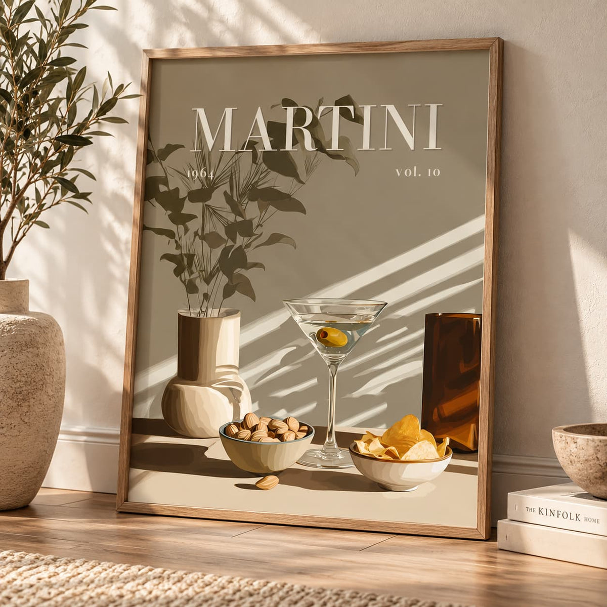 A framed art print leaning against a wall on a wooden floor, featuring a minimalist Martini glass, bowls of snacks, and the word 'MARTINI' in large white letters. The print has a warm beige background with a light wood frame and is styled in a sunlit room with a plant and books nearby.