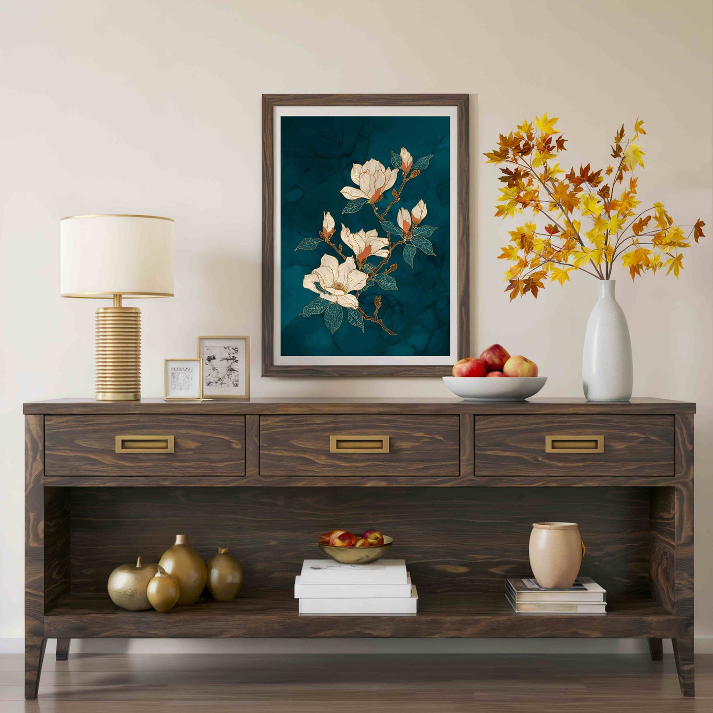 A framed floral art print featuring white magnolia blossoms on a dark teal background, displayed on a dark wood console table in a styled room setting with a lamp, books, and a vase of autumn leaves.