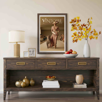 A dark wood console table styled in a living room setting, featuring a framed 'Vintoure' magazine cover print on the wall above it. The table is decorated with a table lamp, picture frames, a vase of autumn leaves, a bowl of apples, and decorative gold-toned ornaments.