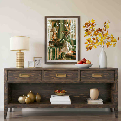 A dark brown wooden console table styled in a living room setting, featuring three drawers with brass handles, an open lower shelf, and decorative items including a lamp, framed art, books, and a white vase with autumn-colored leaves.