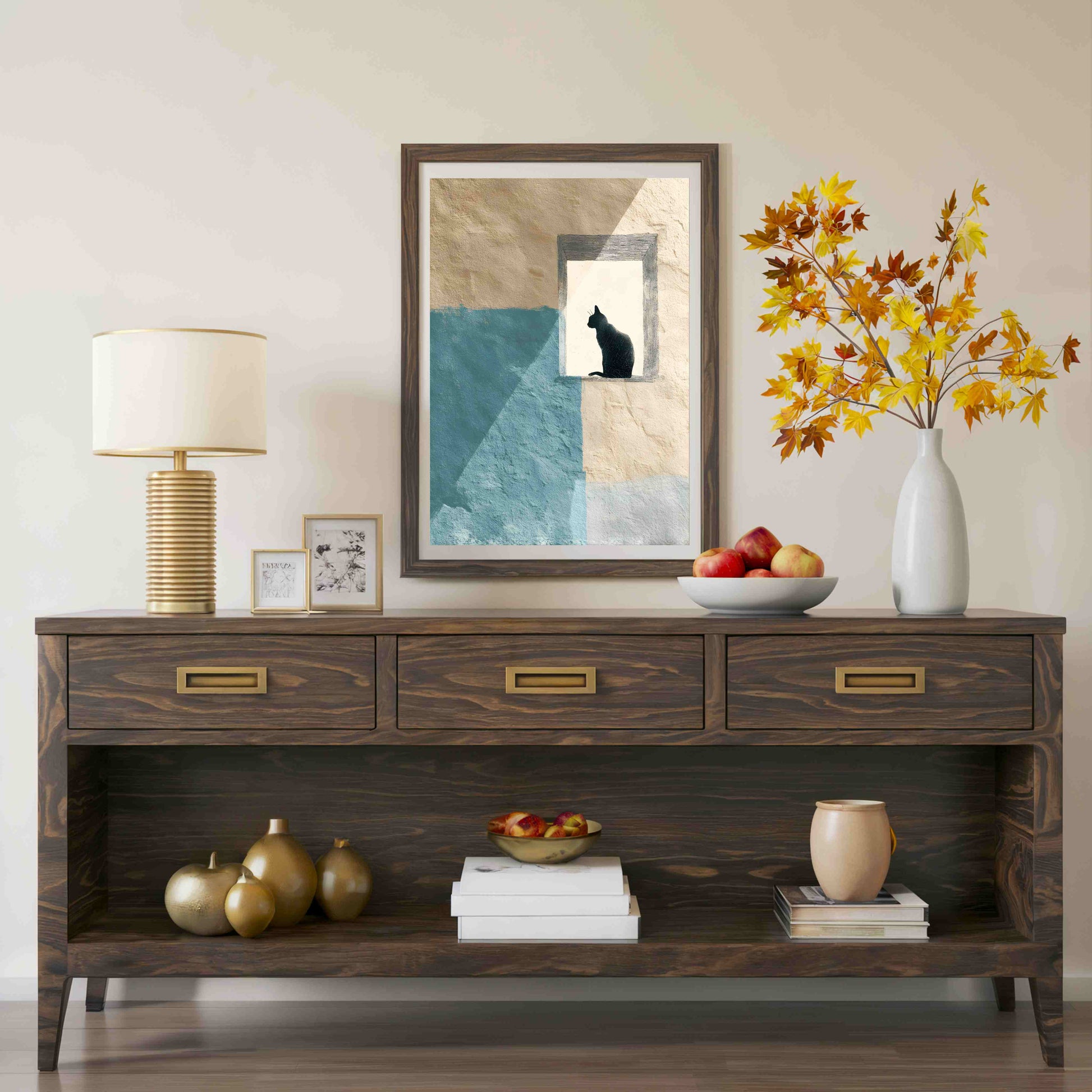 A dark brown wooden console table styled with a table lamp, framed art, a white vase with autumn leaves, fruit, and decorative objects against a neutral beige wall.