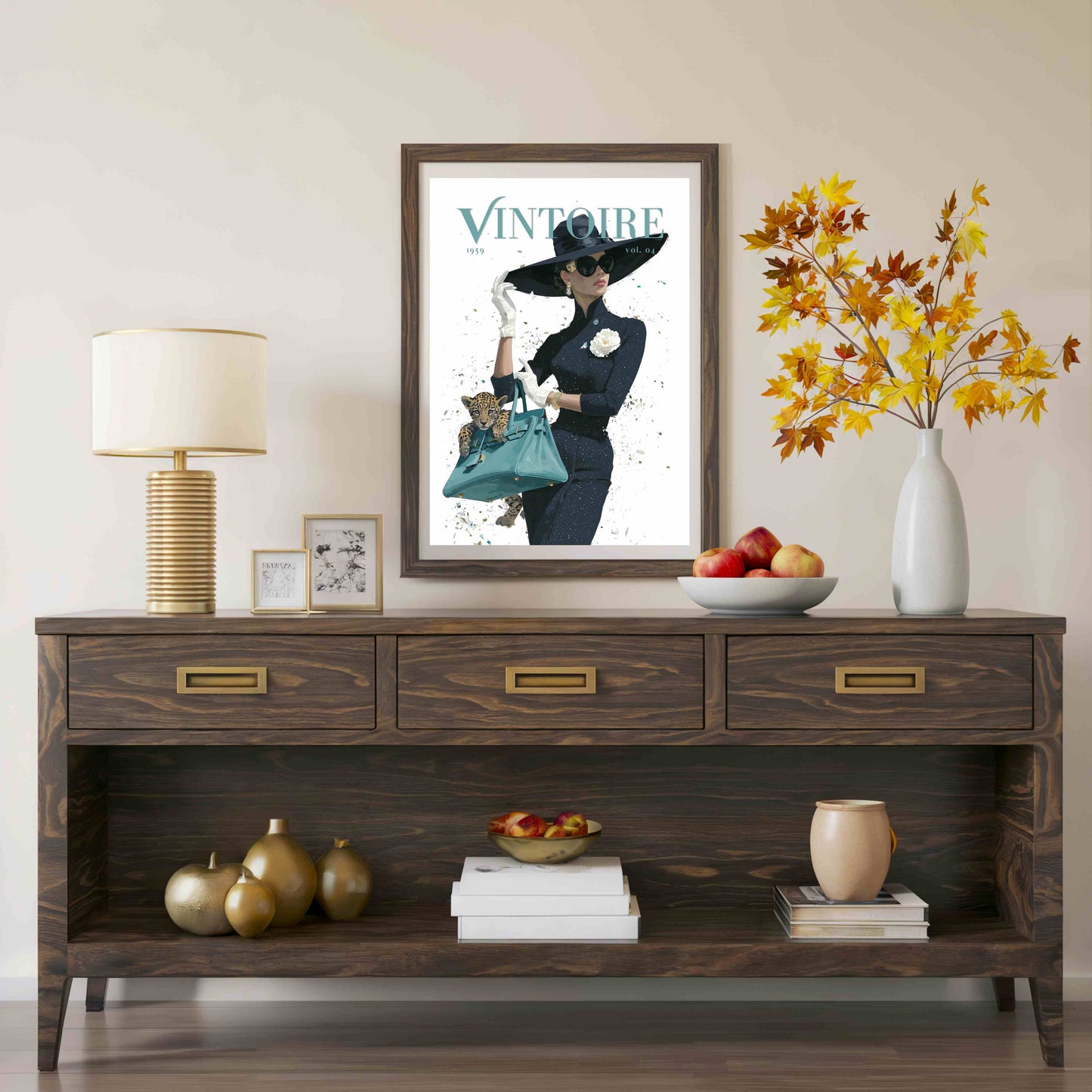 A framed vintage-style poster with the word 'VINTOURE' hangs above a dark wood console table in a styled interior. The room includes a table lamp, small framed art, a bowl of fruit, a vase with autumn leaves, and decorative objects on the table and shelf.